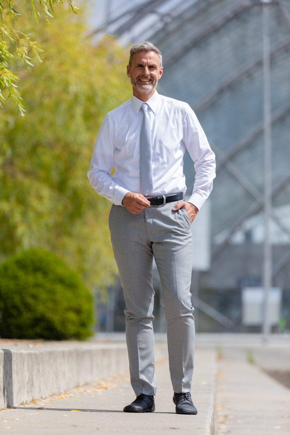 Un hombre con cabello gris y barba está de pie al aire libre en un entorno urbano moderno. Lleva una camisa blanca, pantalones de traje gris claro y elegantes zapatos PEAQ Vegan Black de ZAQQ. Sonríe frente a un fondo de arquitectura de vidrio y verde. Sus zapatos están equipados con un forro de microfibra transpirable para mayor comodidad.