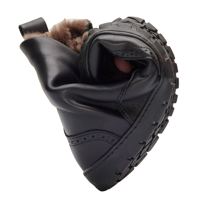 Un flexible QUINTIC Winter Brogue Black de ZAQQ con suela robusta y forro de piel está girado para demostrar su flexibilidad. El zapato, hecho de cuero napa de alta calidad, está posicionado con la piel hacia arriba y la suela muestra su patrón de agarre estructurado.