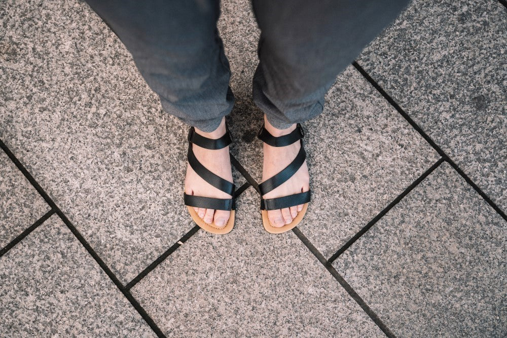Vista de los pies de una persona en las sandalias NIQ Black de ZAQQ de cuero napa de alta calidad, de pie sobre una acera pavimentada con baldosas de piedra. El elegante diseño de correas negras ofrece máxima comodidad y complementa perfectamente los pantalones oscuros.