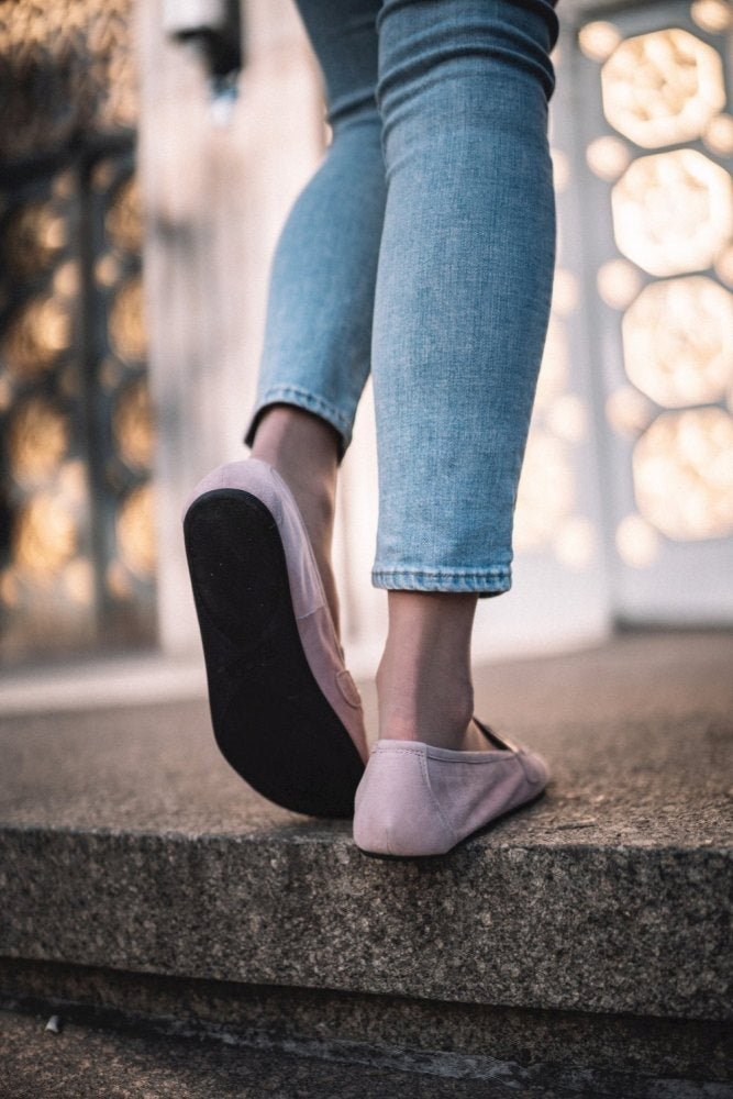 Nahaufnahme einer Person in hellblauer Jeans und rosa ZAQQ QERP Candy Schuhen mit Kautschuksohle, die auf eine Betonfläche tritt. Der Hintergrund ist leicht unscharf und zeigt eine gemusterte Wand.