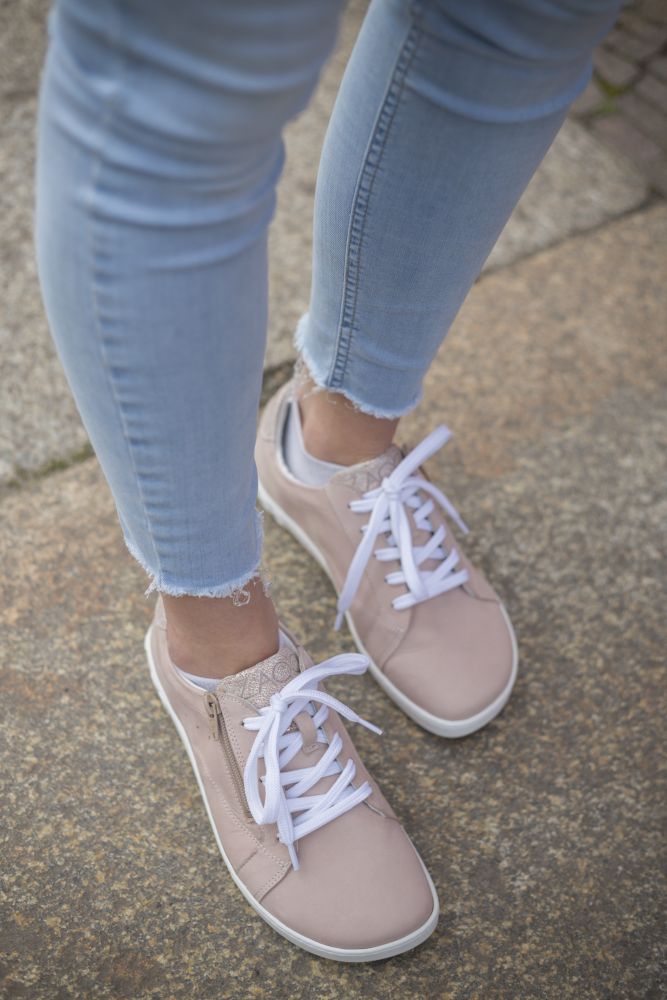 Una persona con jeans azul claro y los elegantes ZAQQ QOMFY Wide Rosa Sneakers con cordones blancos pasea con confianza sobre el pavimento de piedra.
