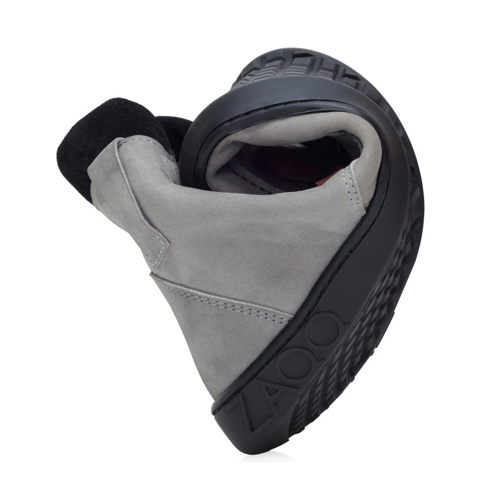 El zapato ROQA Grey está curvado para demostrar su flexibilidad y ofrece comodidad flexible. Cuenta con una suela oscura y una parte superior gris claro. El nombre de la marca ZAQQ está grabado en la suela y resalta el diseño y la comodidad del zapato, que recuerda a los zapatos descalzos.