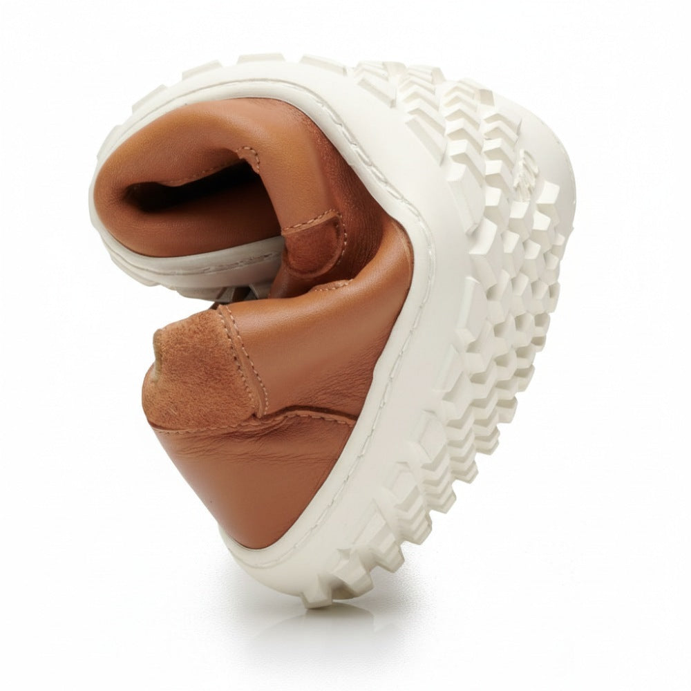 Der SHOQQ-Cognac von ZAQQ, ein hellbrauner Ledersneaker mit einer dicken weißen Gummisohle, ist in der Mitte gefaltet, um seine Flexibilität und seinen Komfort hervorzuheben. Die Sohle weist ein ausgeprägtes, robustes Profilmuster auf, das dem eines Barfußschuhs ähnelt. Der Schuh steht vor einem schlichten weißen Hintergrund.