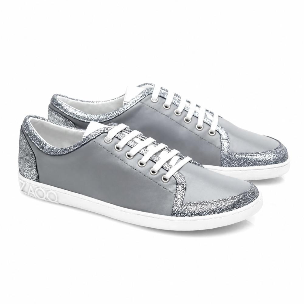 Las zapatillas TIQQ Grey Silver de ZAQQ destacan por su diseño sencillo de napa suave con cordones blancos y acentos de brillo plateado, complementadas con suelas blancas y un discreto logo en el talón, ofreciendo la mezcla perfecta de estilo y comodidad para cualquier ocasión.