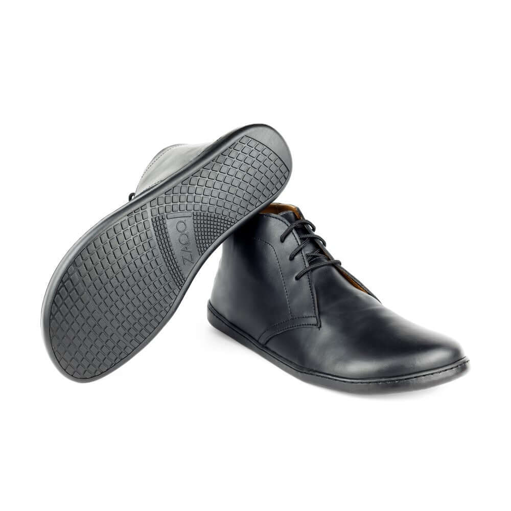 Schwarze Lederstiefeletten auf weißem Hintergrund. Ein Stiefel steht aufrecht und hat eine glatte, polierte Oberfläche mit Schnürsenkeln, während der andere mit der Sohle nach oben steht und ein strukturiertes Muster für Halt aufweist. Der Markenname ist in die Sohle eingeprägt.