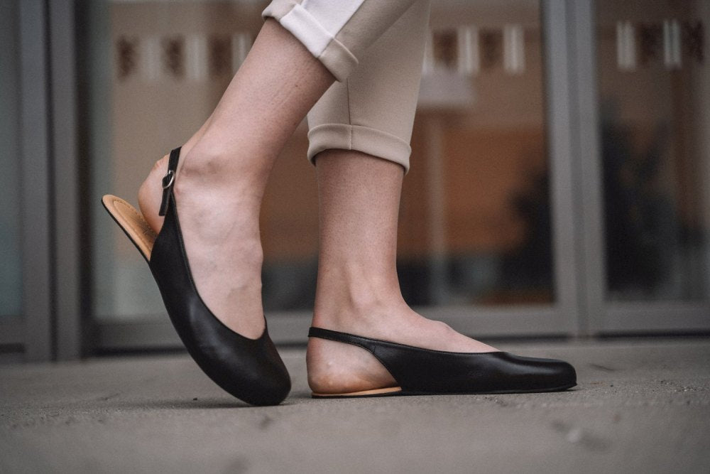 Nahaufnahme einer Person, die elegante PINQ Black Slingback-Schuhe von ZAQQ trägt, die den Komfort eines Barfußschuhs bieten, kombiniert mit beigefarbenen, hochgekrempelten Hosen. Der Fokus liegt auf ihren Füßen, während sie auf einer Betonoberfläche stehen, mit einem verschwommenen Gebäude im Hintergrund.
