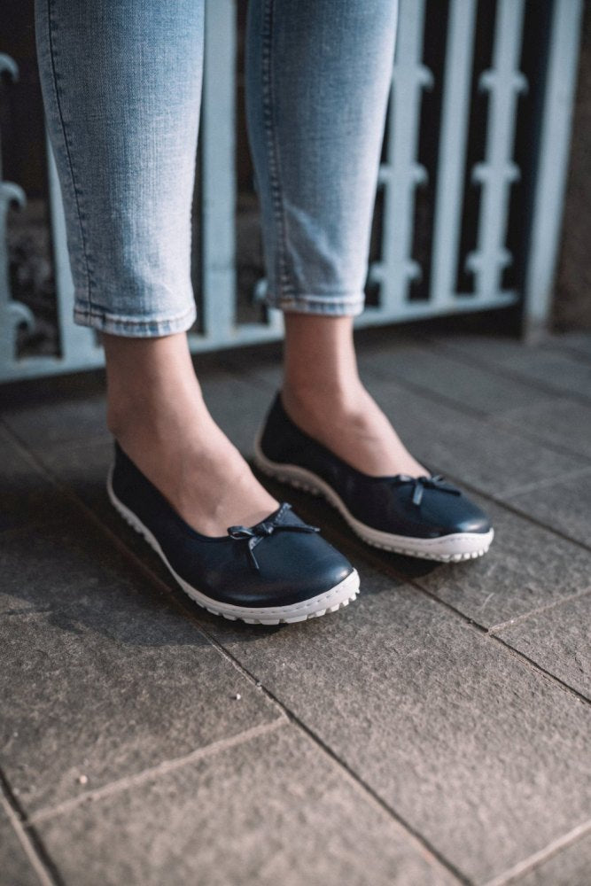 Eine Person in dunkelblauen SUQAR-Jeans und schwarzen Slippern mit strahlend weißen Sohlen steht auf einem gefliesten Boden. Im Hintergrund ist ein dekoratives Metalltor zu sehen.